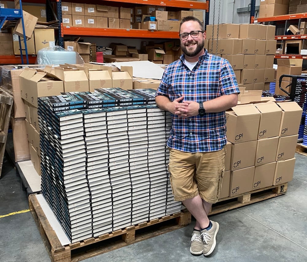 Me (bearded white man in checked shirt, shorts and glasses) beside a pile of several hundred copies of my books in a warehouse.