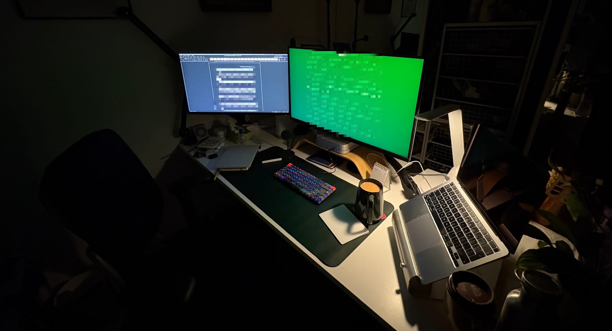 A dimly lit desk with two monitors, a glowing keyboard, a large cup of tea and a laptop off to one side